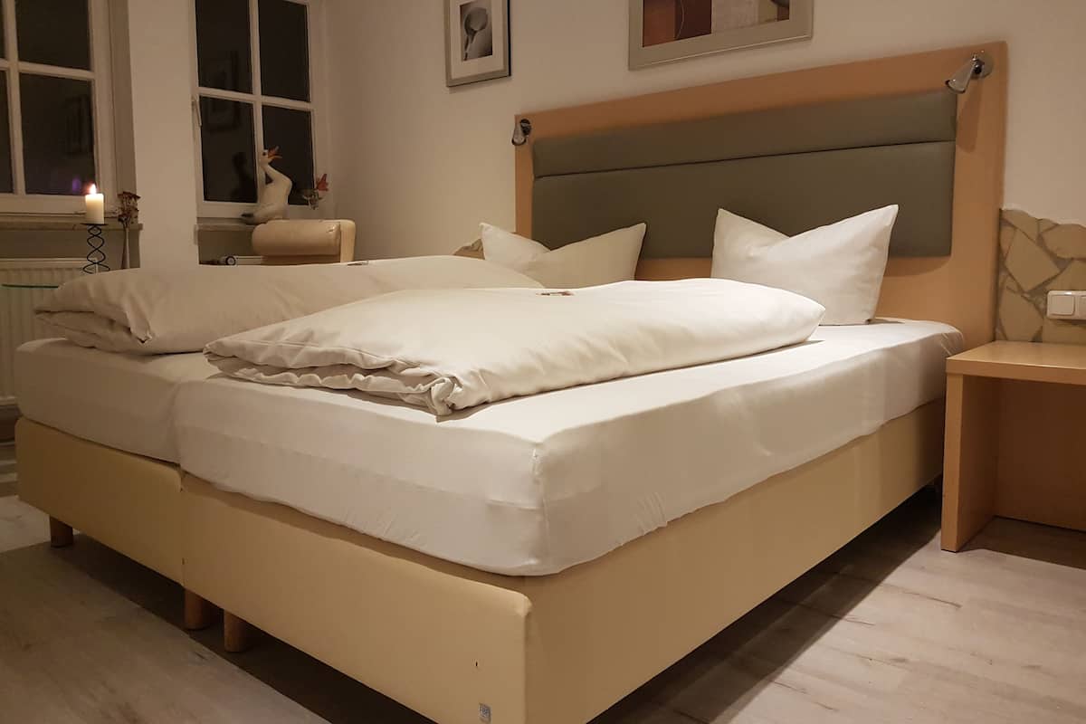 Una cama bien hecha con sábanas y almohadas blancas, una mesita de noche con una lámpara, y una ventana con una vela encendida.