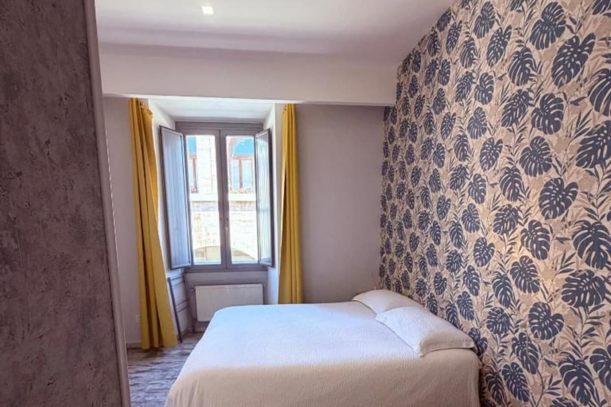 A bedroom with a bed, a small stool, a window with yellow curtains, and a patterned wall.