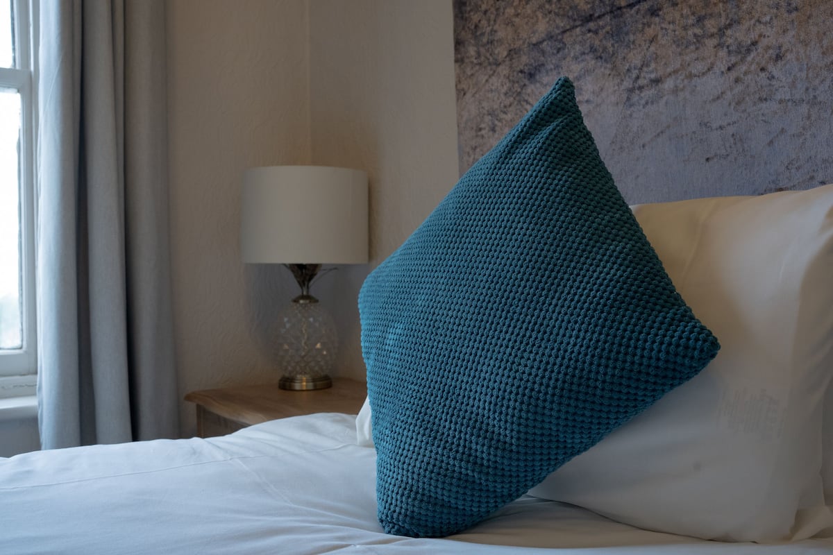 A neatly made bed with a teal pillow and a white pillow, a bedside lamp, and a window with curtains.
