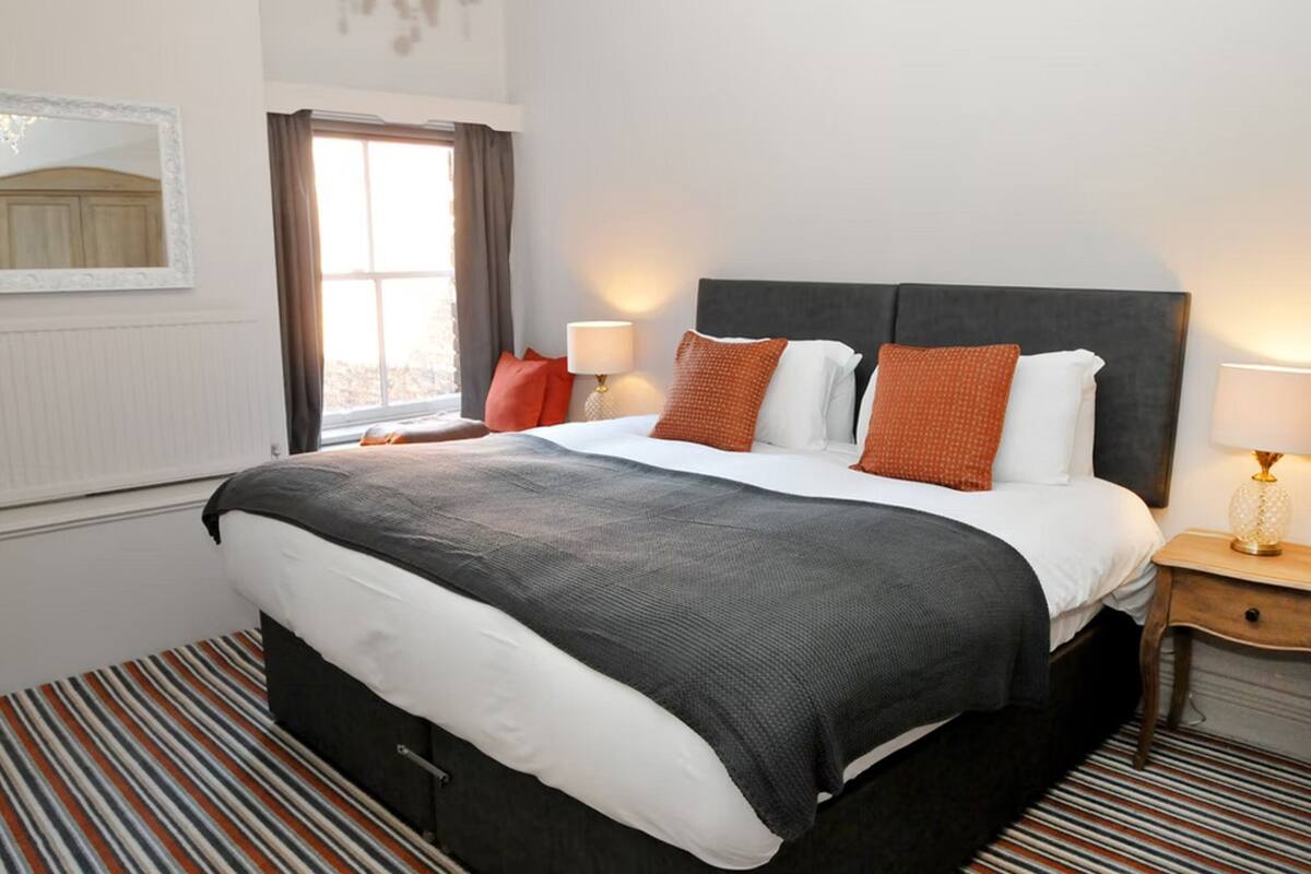 A neatly made bed with a dark headboard, white bedding, and orange pillows. There is a window with curtains, a lamp on the bedside table, and a striped rug on the floor.