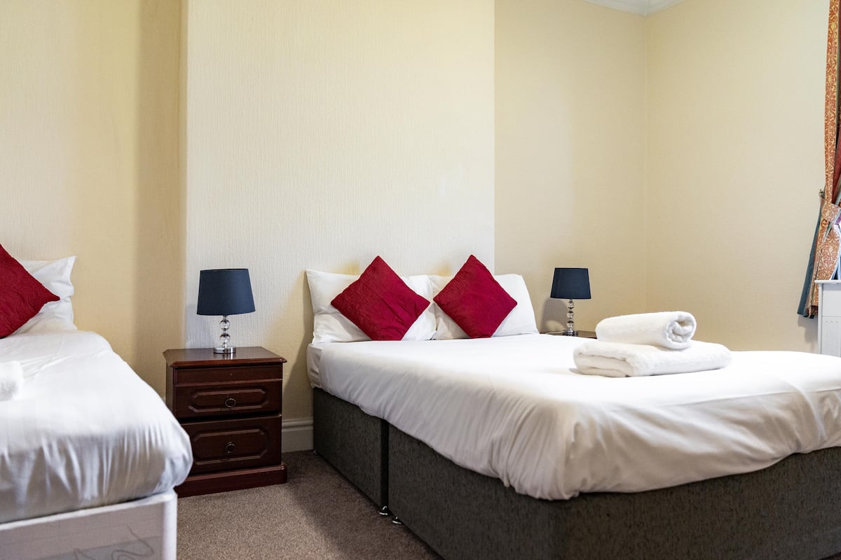 A hotel room with two beds, each with white linens and red pillows, a small wooden nightstand with a lamp, and a neatly folded towel on the larger bed.