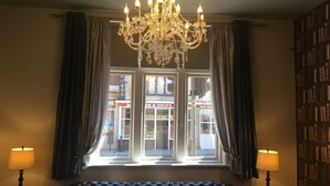 A room with a black leather sofa, a chandelier, two lamps, a bookshelf, and a view of a street with shops. - The Royal Hotel Crewe, A BRILLIANT Collection (Crewe)