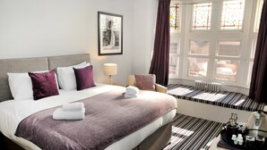 A hotel room with a large bed, a bedside table with a tray of toiletries, a window with curtains, and a radiator. - The Royal Hotel Crewe, A BRILLIANT Collection (Crewe)