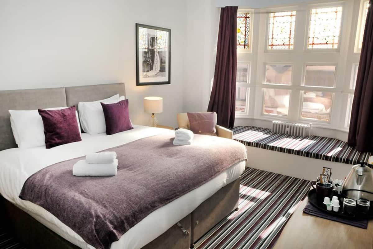 A hotel room with a large bed, a bedside table with a tray of toiletries, a window with curtains, and a radiator.