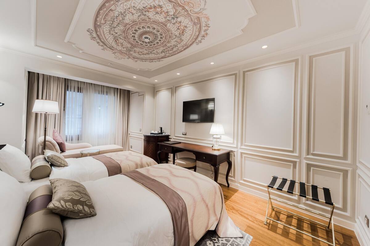 A hotel room with two beds, a desk, a television, and a ceiling with intricate design.
