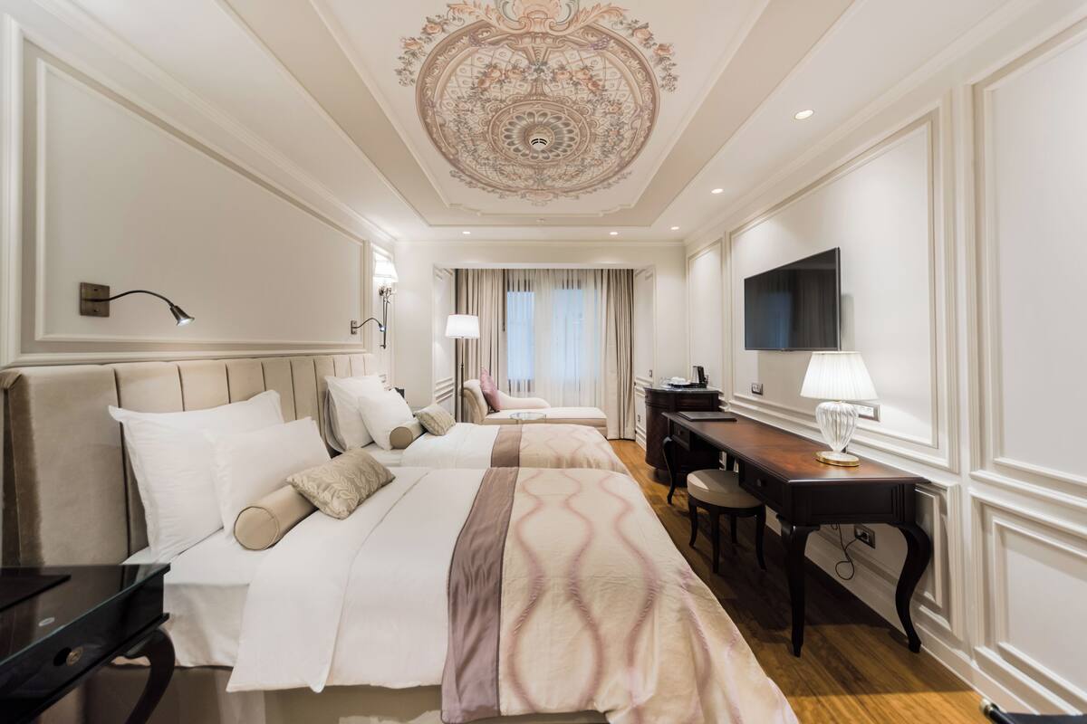 A hotel room with two beds, a desk, a chair, a TV, and a decorative ceiling.
