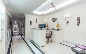 A hotel lobby with a long corridor, a reception desk with a computer, a chair, and wall-mounted artwork.