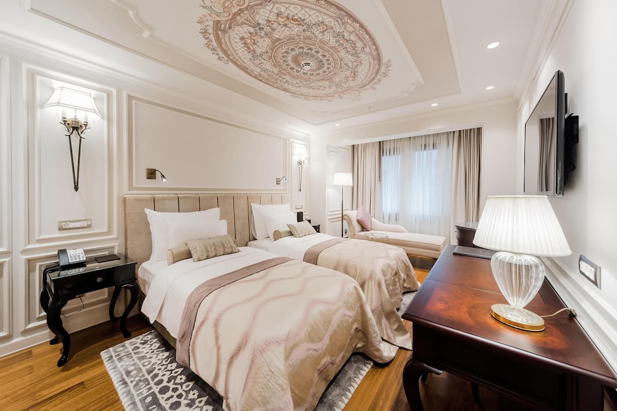 A hotel room with two beds, a wooden desk, a lamp, and a ceiling with intricate design.