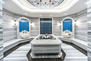 A luxurious bathroom with marble walls, two sinks, a central heated stone bench, and a decorative ceiling.