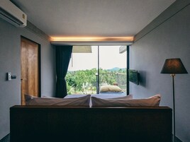 A hotel room with a bed, a window with a view of greenery, a door, and a wall-mounted TV.