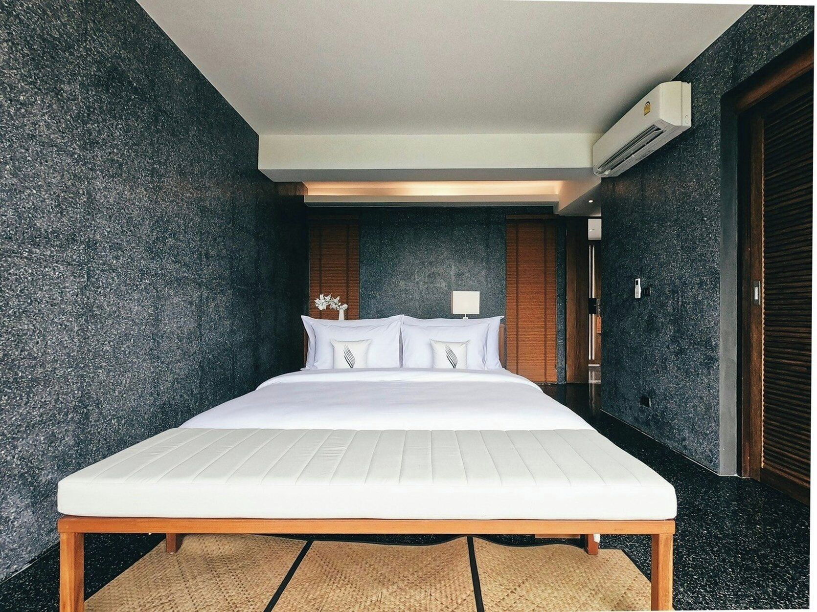 Una habitación de hotel moderna con una cama grande, paredes texturizadas oscuras, piso de madera y un equipo de aire acondicionado.