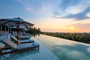 Une piscine à débordement bordée de chaises longues et d’ombrelles, entourée d’une végétation luxuriante et offrant une vue imprenable sur le coucher de soleil.