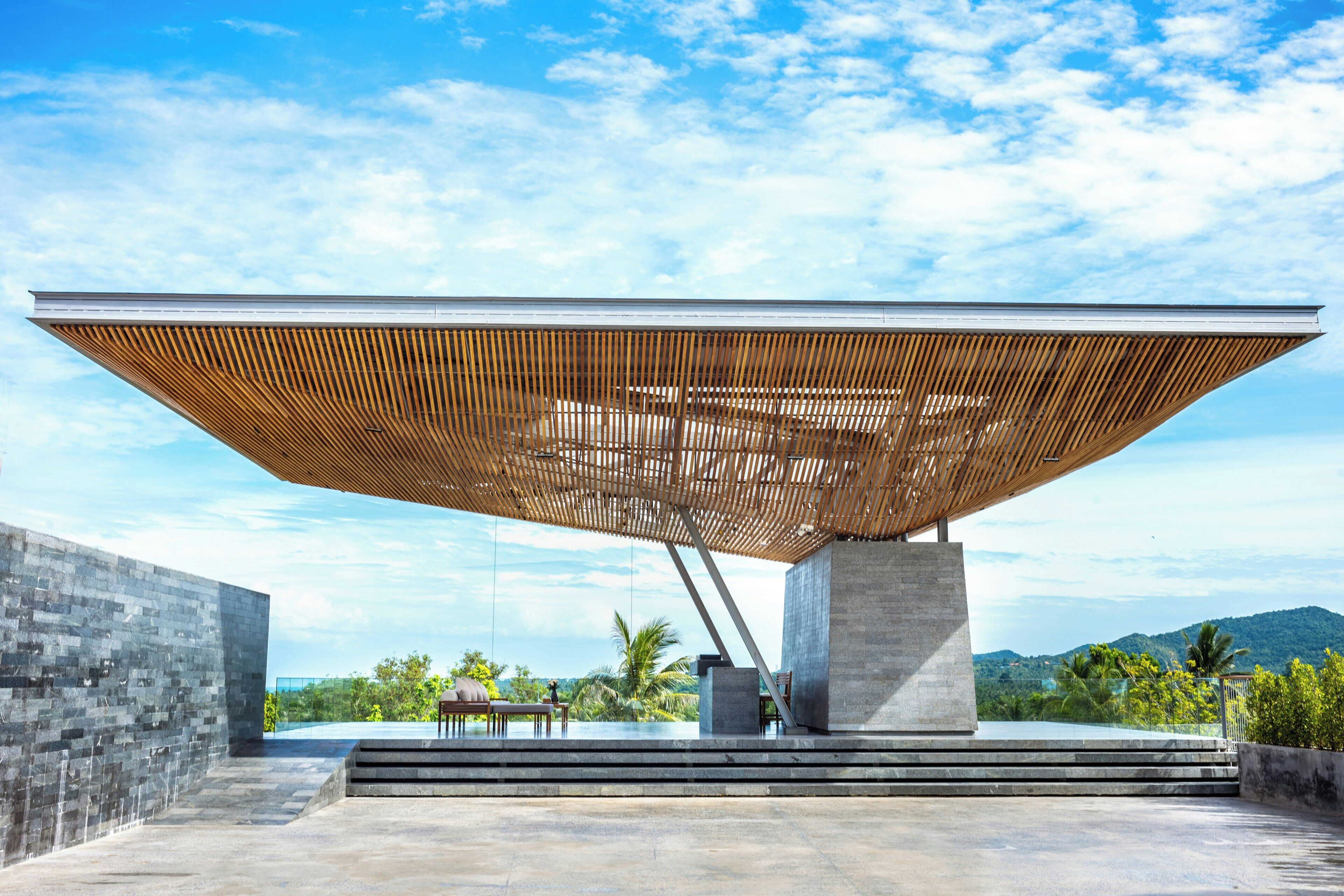 A modern outdoor structure with a slatted roof, a concrete base, and a view of a mountainous landscape.
