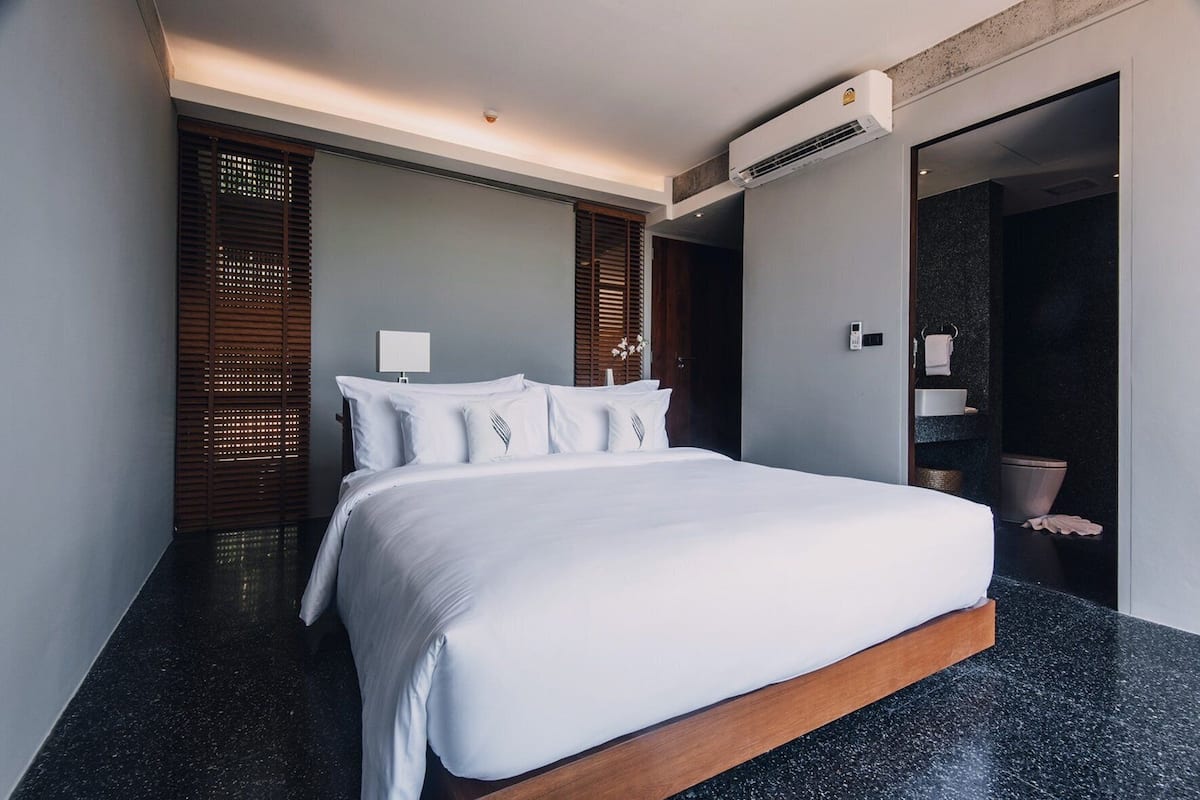 A modern hotel room with a large bed, a bedside table, a wall-mounted air conditioner, and a bathroom visible through an open door.