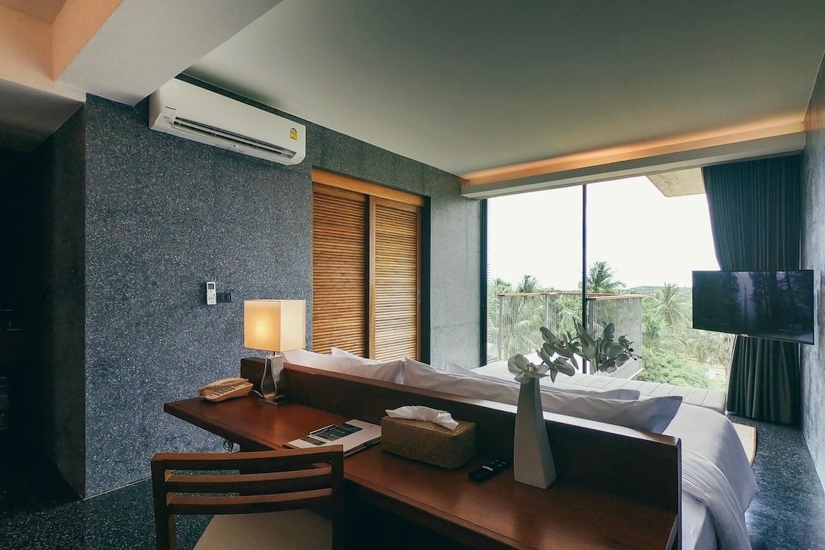 A hotel room with a bed, a desk with a chair, a TV, an air conditioner, a window with a view of palm trees, and a bedside table with a lamp and a tissue box.