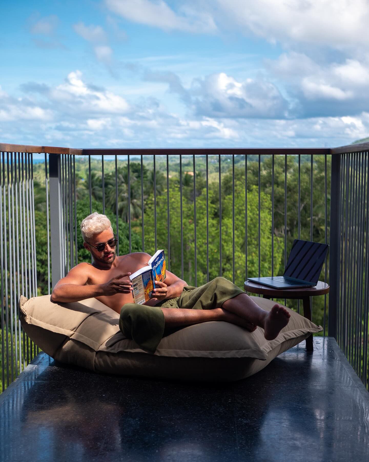 A person lying on a cushioned seat on a balcony with a laptop and a book, overlooking a lush green landscape.