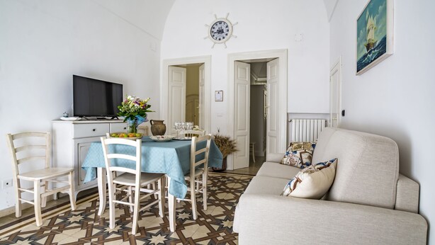 A living room with a sofa, a dining table with chairs, a TV, and a clock on the wall. - Domus Neptuni (Monopoli)