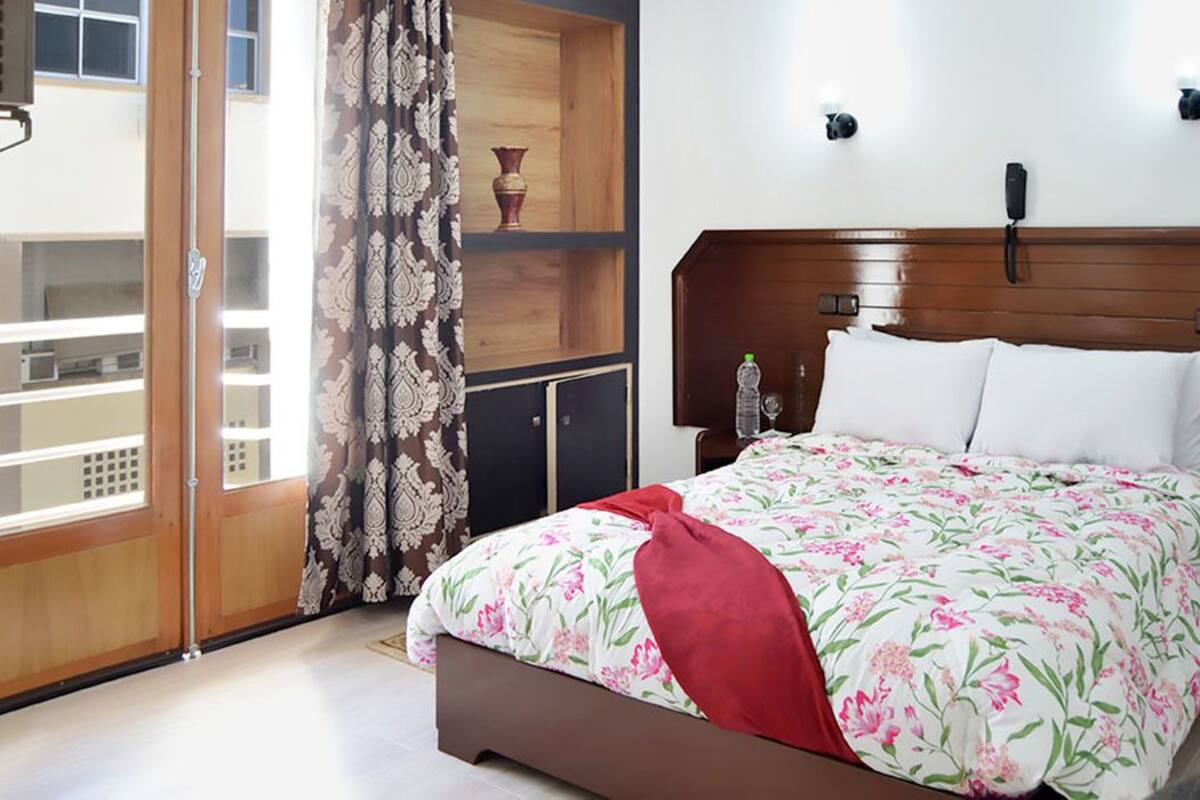 Une chambre à coucher avec un lit, une tête de lit en bois, une table de chevet, un téléphone et une fenêtre avec un rideau.