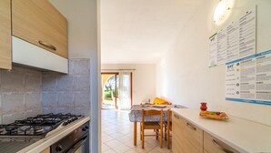 A kitchen with a gas stove, oven, and wooden cabinets. There is a dining table with chairs and a window in the background. - Residenza Lu Fraili (San Teodoro)