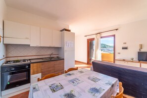 Cocina compacta con estufa, horno y armarios de madera. Zona de comedor con mesa y sillas, y vista al exterior a través de una puerta.