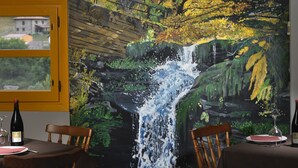 Un comedor con un mural que muestra una cascada y follaje otoñal. Hay dos mesas de madera con sillas, un ventanal con marco amarillo y una botella de vino sobre la mesa.