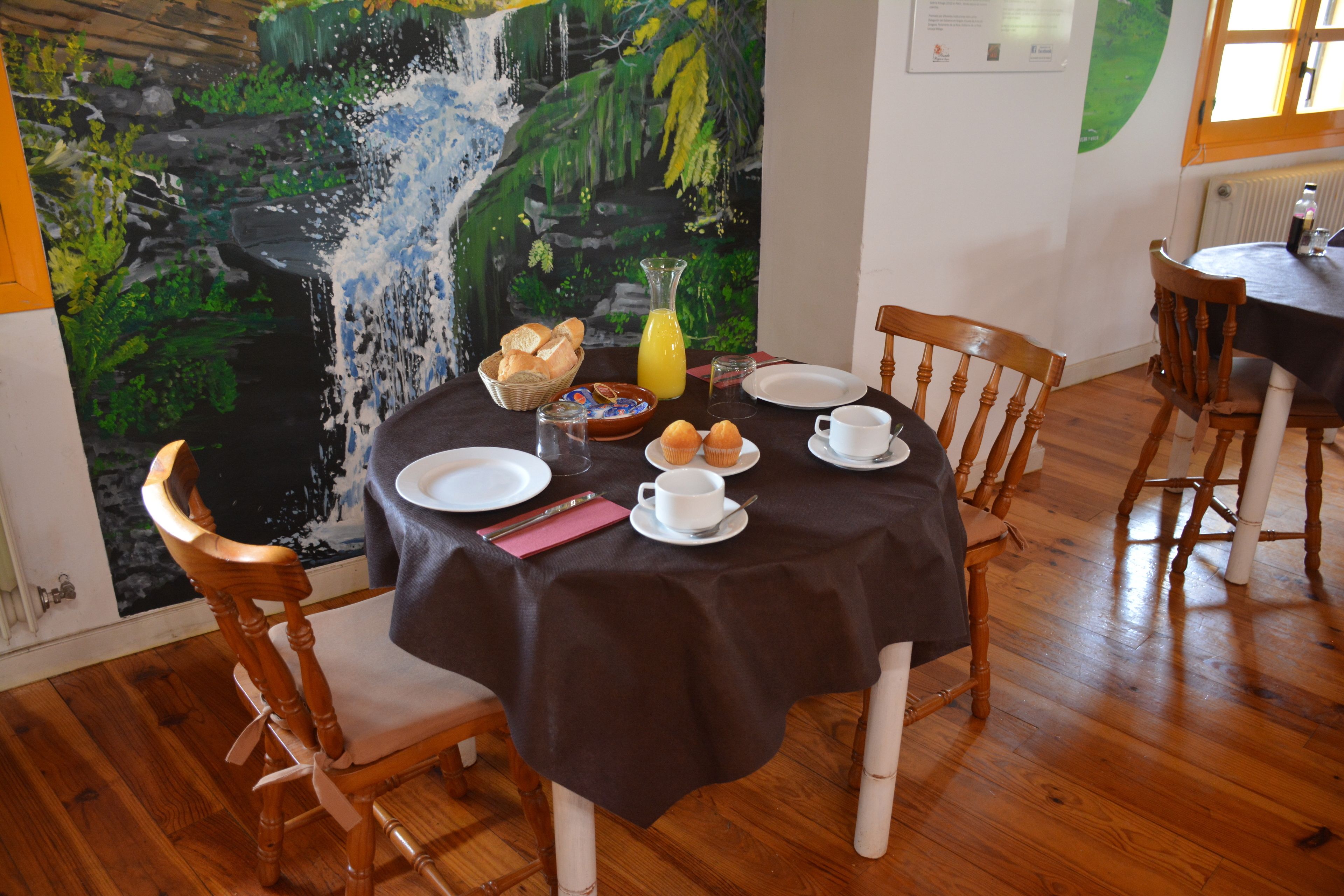 Une table à manger dressée avec des assiettes, des tasses et un panier de pain, et une peinture représentant une cascade en arrière-plan.