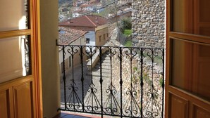 Una puerta de madera abierta que da a un balcón con barandilla de hierro forjado, con vistas a un edificio de piedra y una colina con casas.