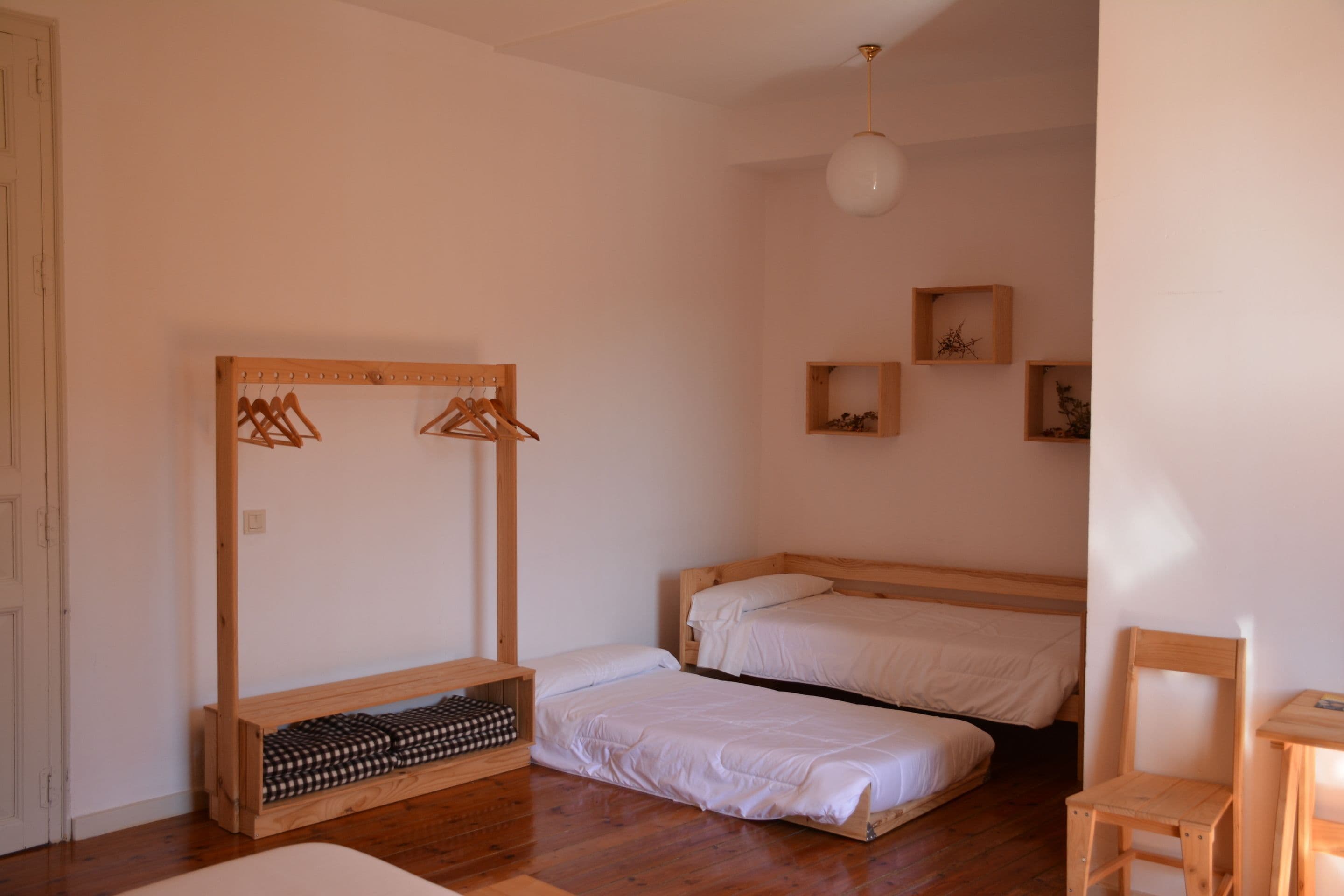 Una habitación con dos camas, un banco de madera, una silla y un estante de madera con ropa colgada.