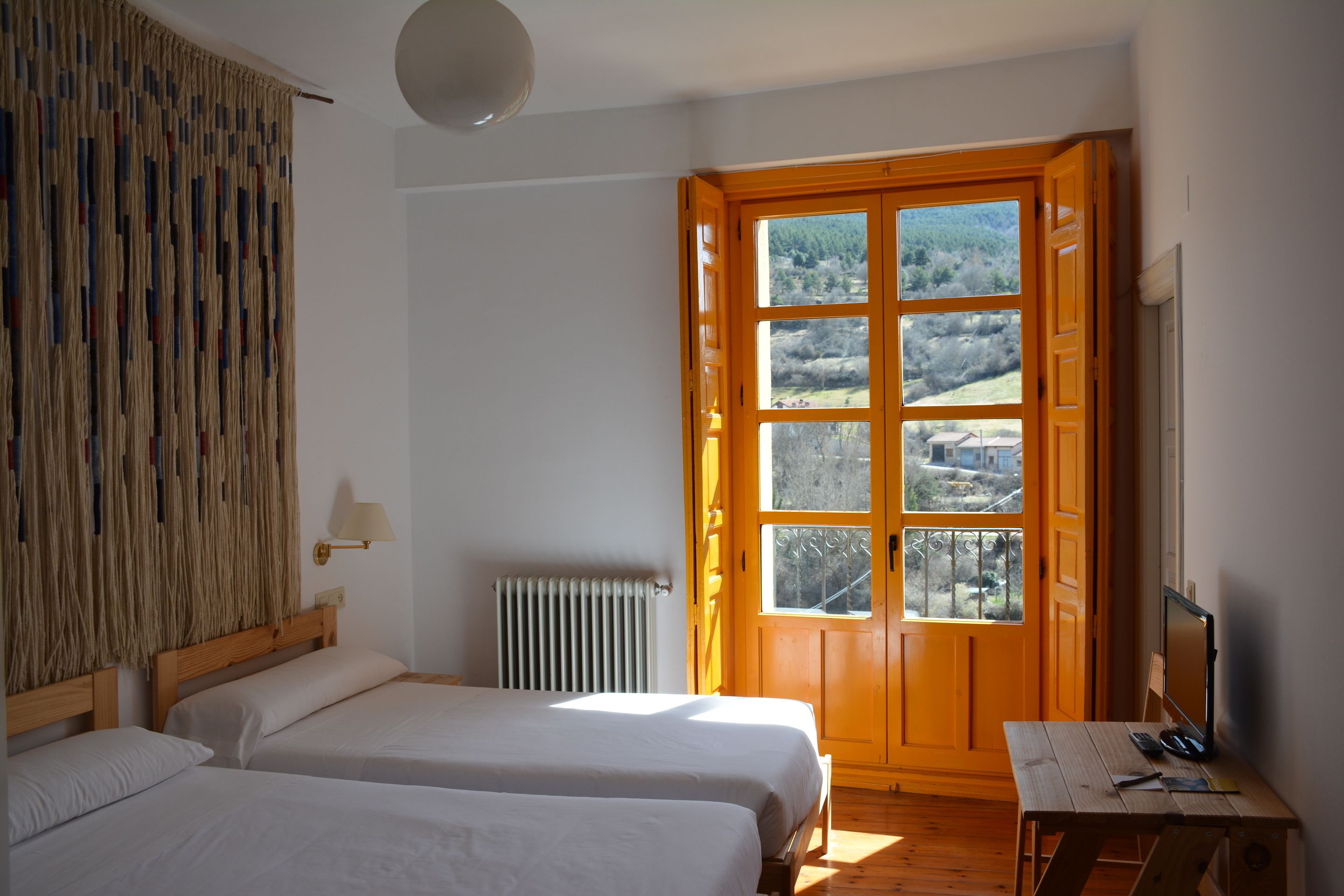 Una habitación con dos camas, un escritorio de madera, un ventanal con vistas al paisaje y un radiador.