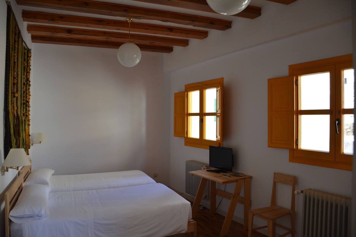 A room with a wooden ceiling, a bed with white bedding, a desk with a computer, and a chair.