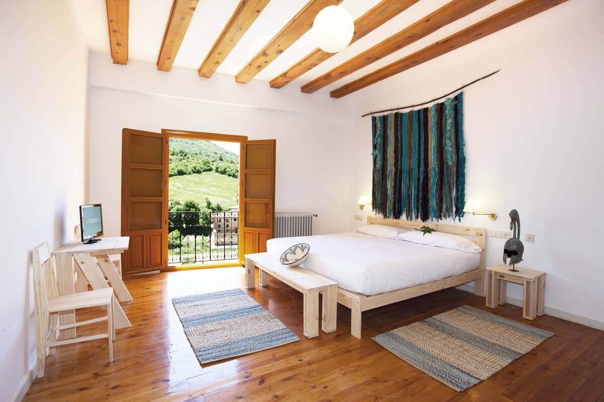 A bedroom with a wooden ceiling, a bed, a desk, and a view of a green landscape through an open door.