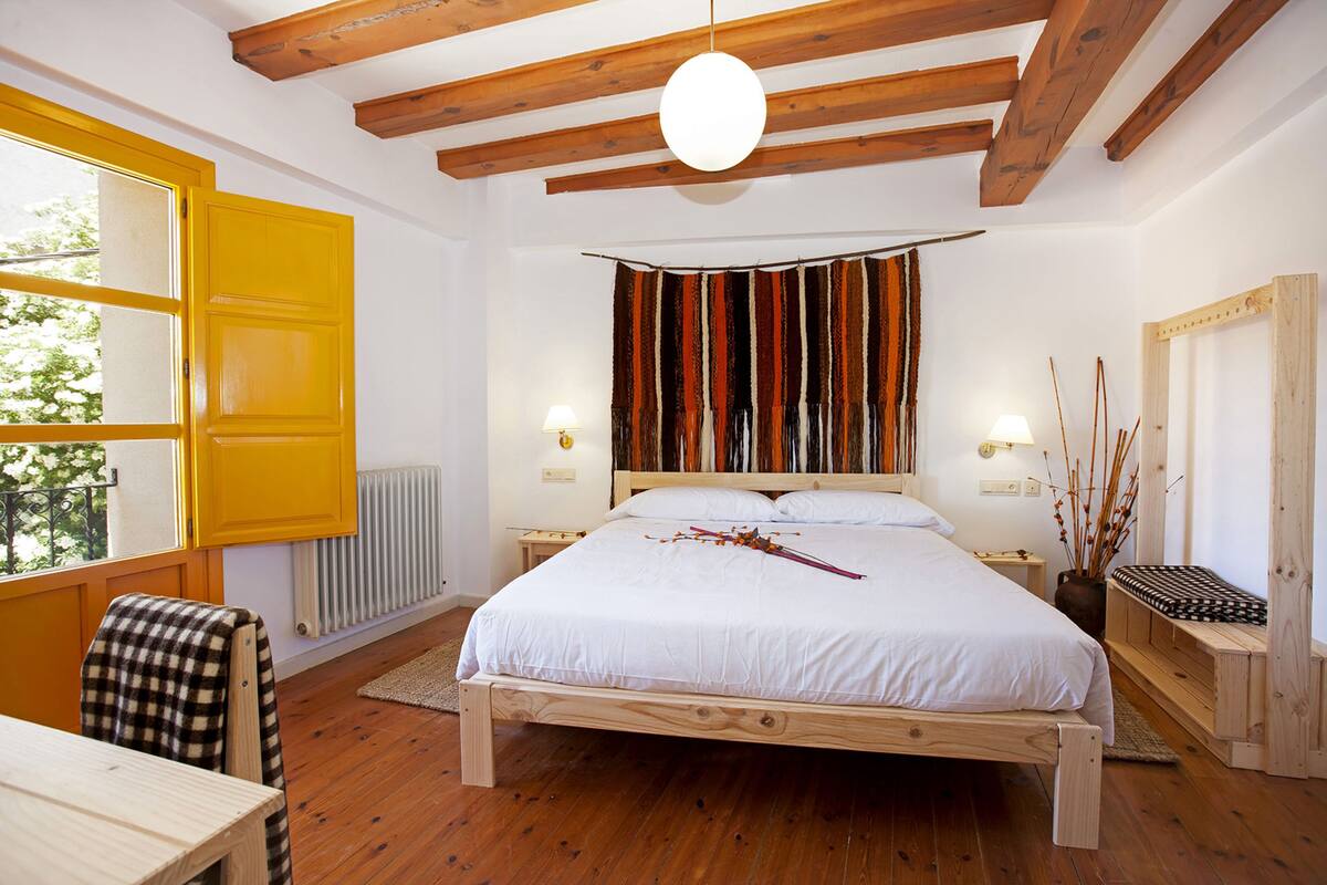 A bedroom with a wooden bed, a window with yellow shutters, a radiator, and a wooden ceiling.