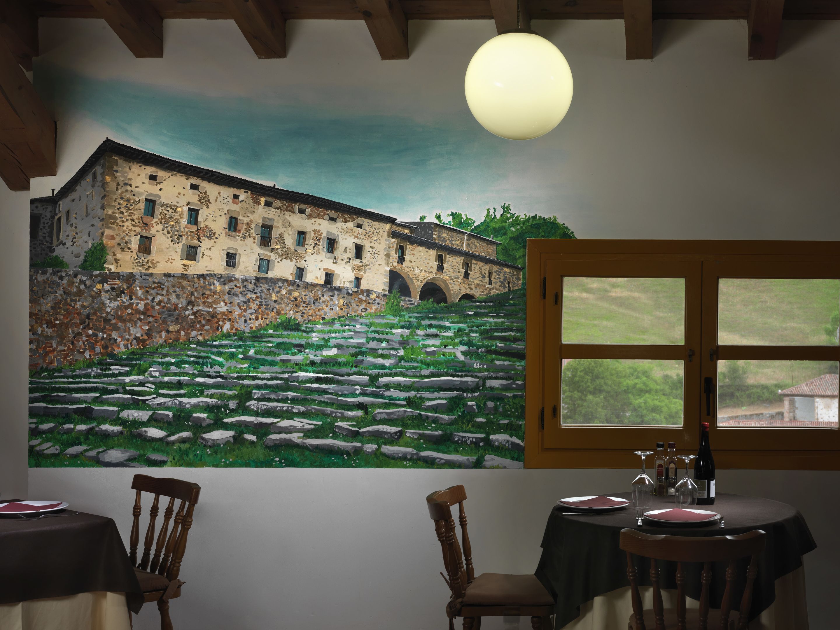 A dining area with a mural of a building and landscape, wooden chairs, a table with a black tablecloth, and a window with a view of greenery.