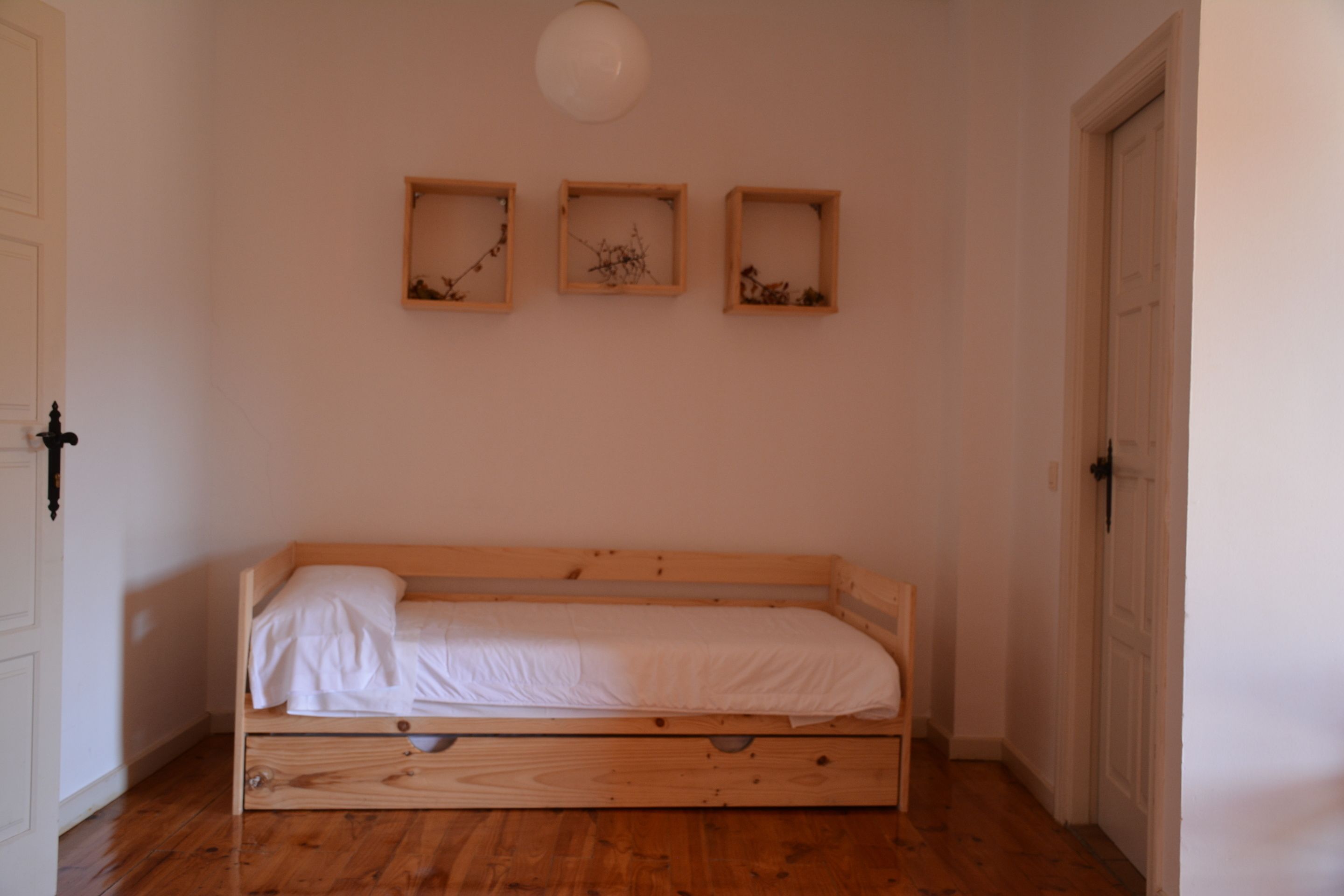 A small, single-bed room with wooden flooring, a white door, and three wall-mounted picture frames.