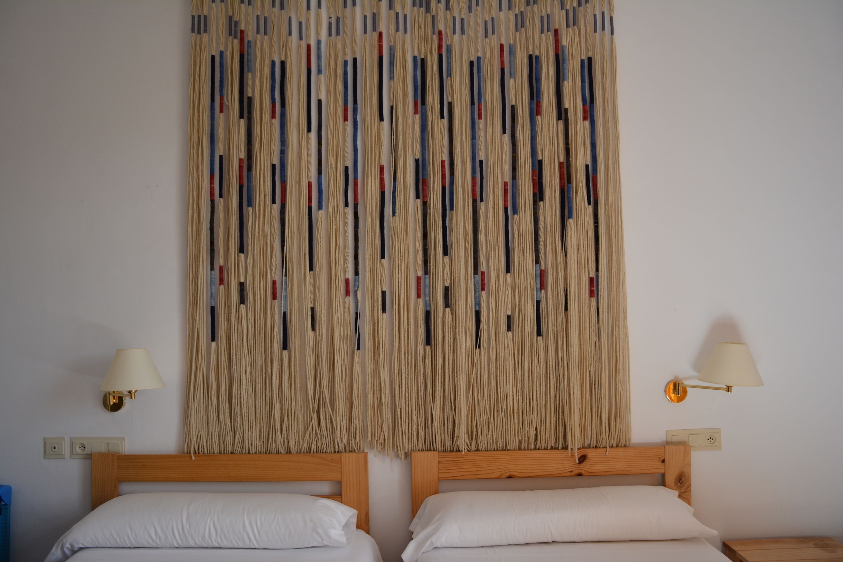 Quarto com duas camas, um painel decorativo na parede e duas luminárias fixadas na parede.