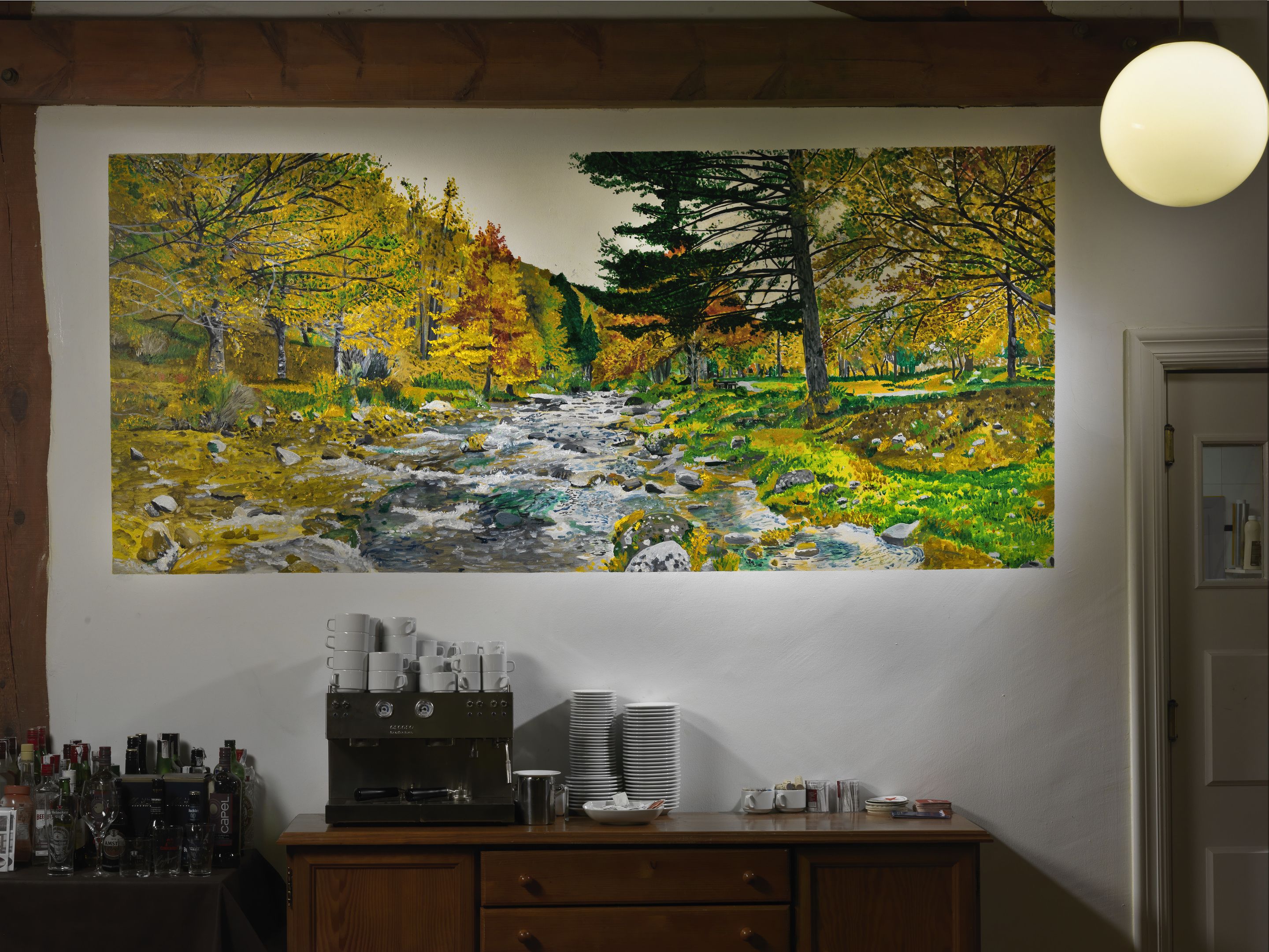 A painting of a river in autumn foliage on a wall above a wooden counter with coffee-making equipment.