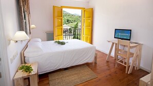 Un dormitorio con cama, escritorio con computadora, ventana con puerta abierta y vista a un paisaje verde.