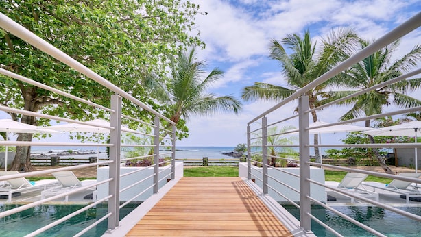 Una terraza de madera que da a una piscina con barandillas blancas y sillas de descanso, con vistas a una playa y al océano.