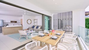 Un comedor moderno con una mesa de madera y sillas de ratán blanco, una cocina al fondo y un balcón con barandilla de vidrio.