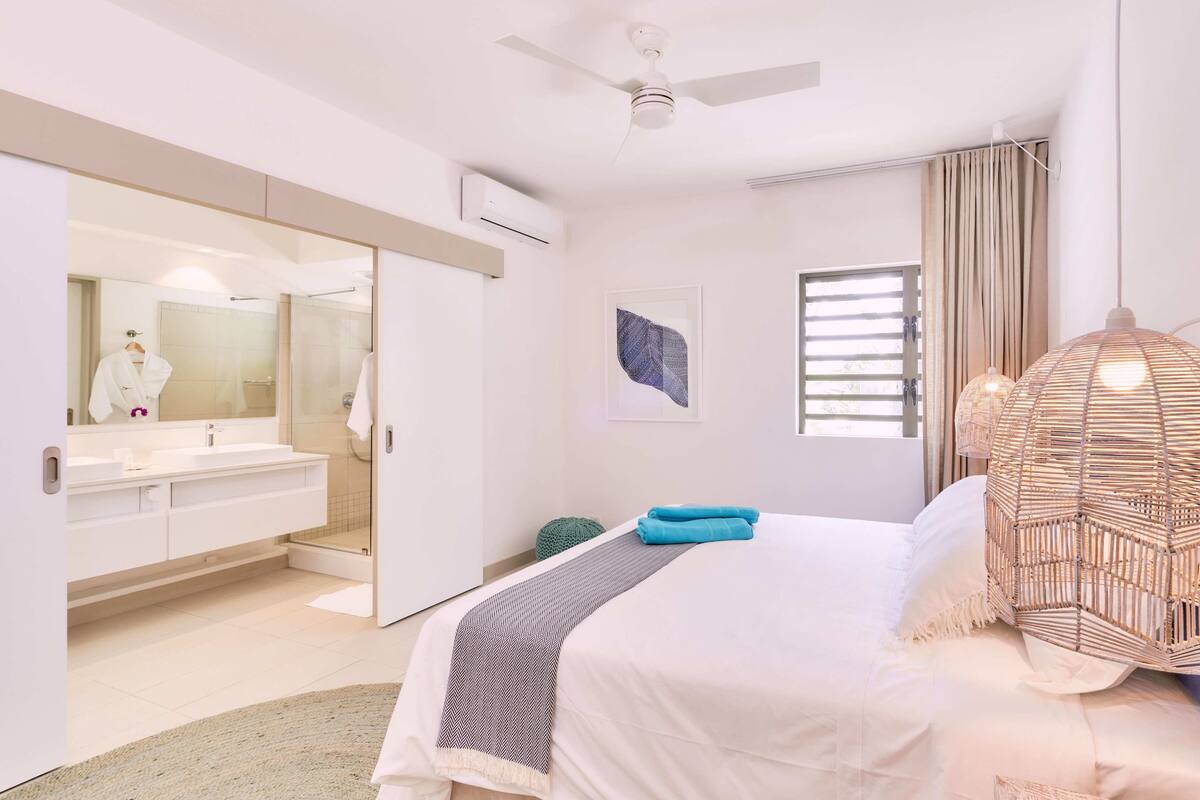 Ein modernes Schlafzimmer mit einem großen Bett, einem begehbaren Kleiderschrank, einem Badezimmer mit großem Spiegel und einem Deckenventilator.