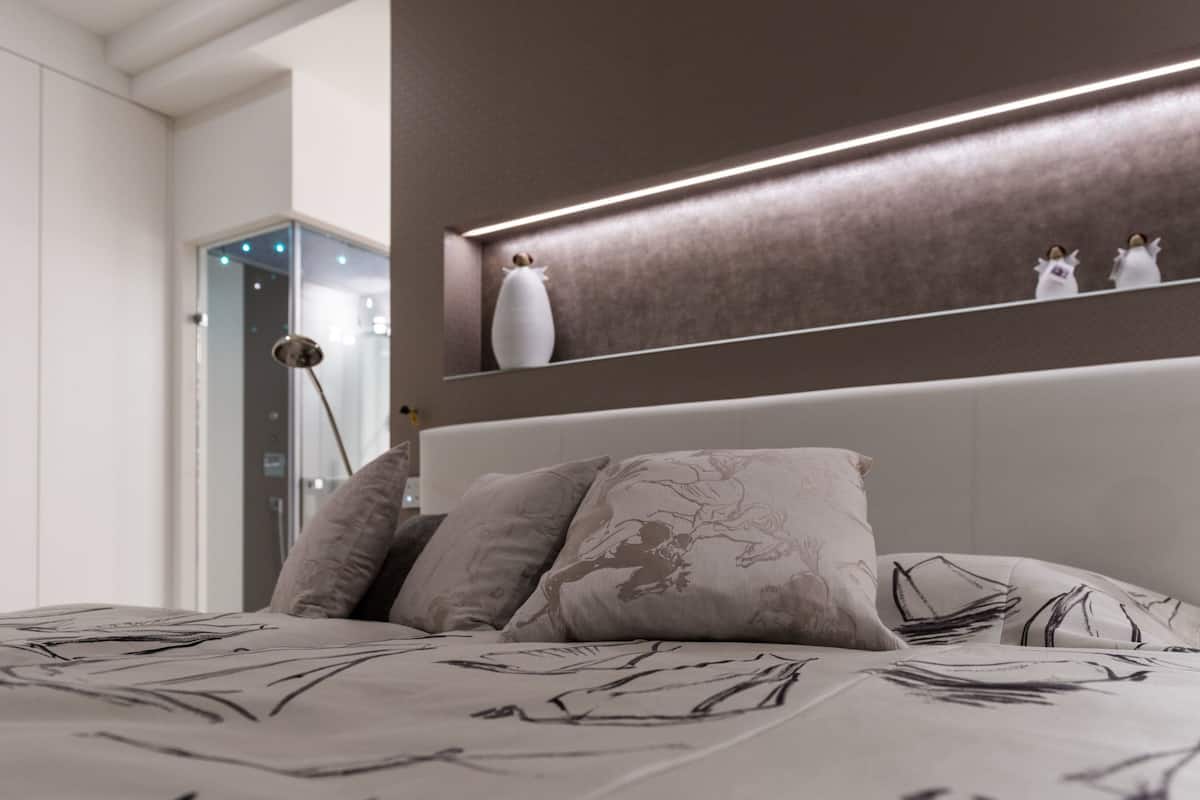 A modern bedroom with a large bed, a headboard with integrated lighting, and a bedside table with decorative items.