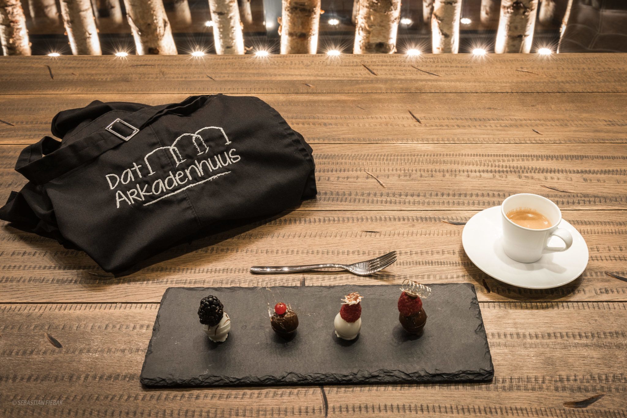 A wooden table with a black bag, a white cup with a saucer, and a slate board with four desserts.