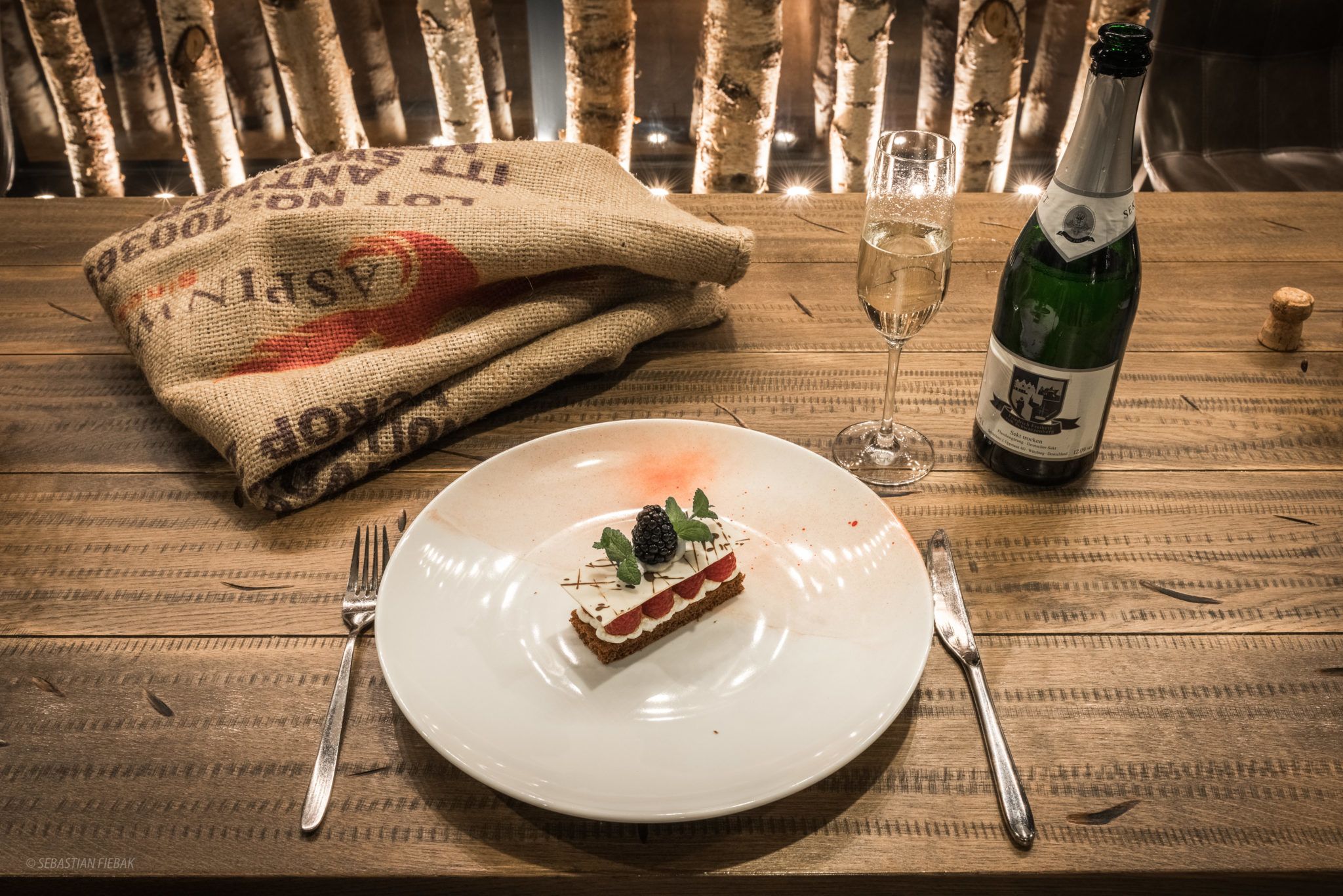 A wooden table with a white plate, a fork, a knife, a bottle of champagne, and a glass.