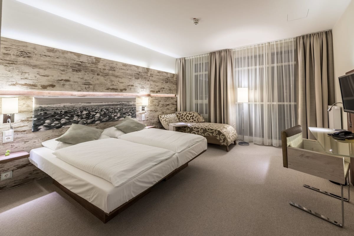 A modern hotel room with a large bed, a seating area with a patterned chair, a television, and a stone wall feature.