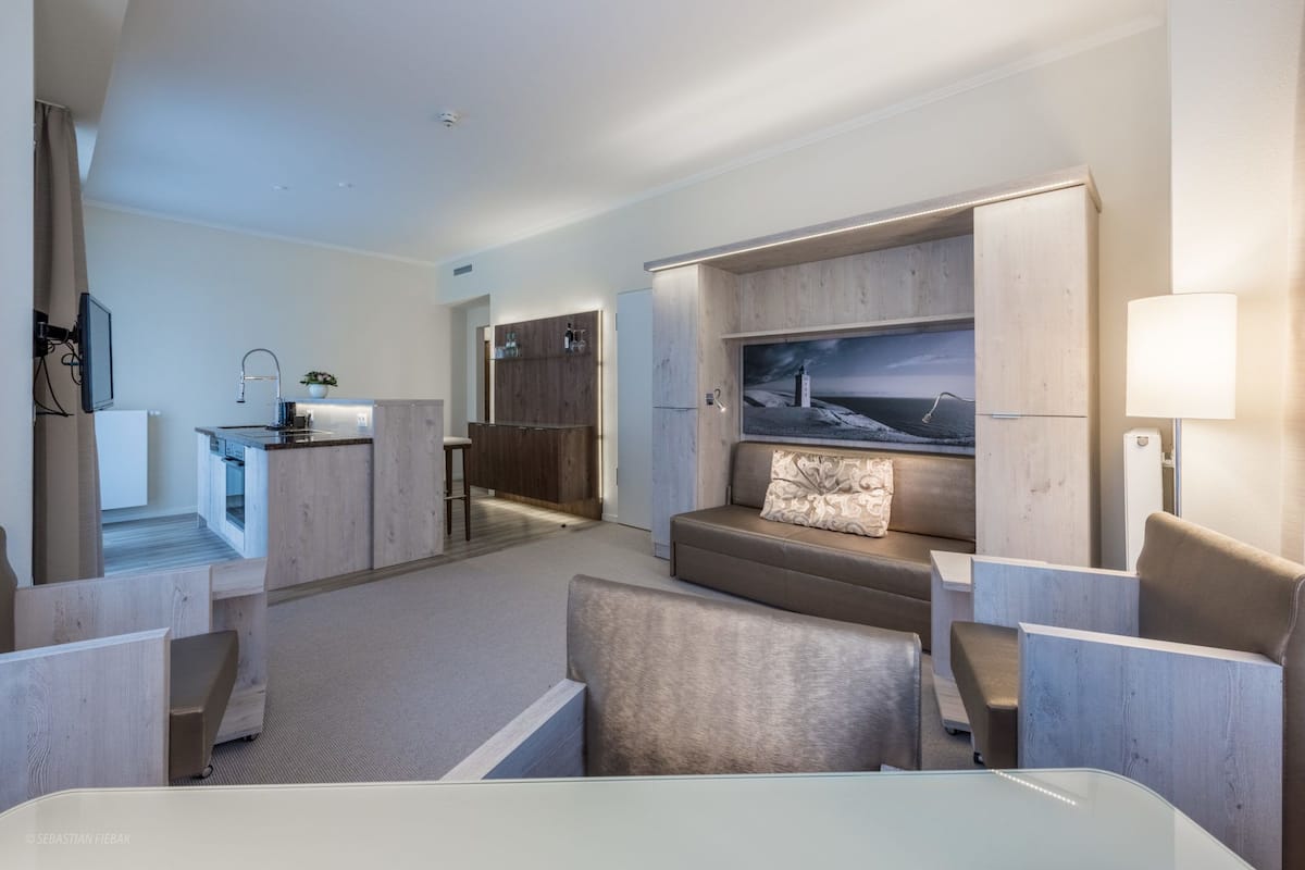 A modern hotel room with a kitchenette, a seating area with a sofa and armchairs, and a large TV mounted on the wall.