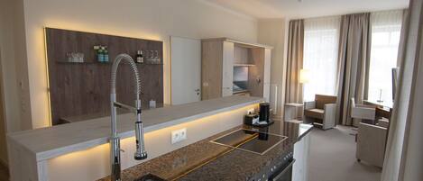 Una cocina moderna con encimera de granito, grifo de acero inoxidable y electrodomésticos integrados. Se aprecia una pared revestida de paneles de madera con estantes sobre el lavabo, y un área de descanso con sofá y mesa al fondo.