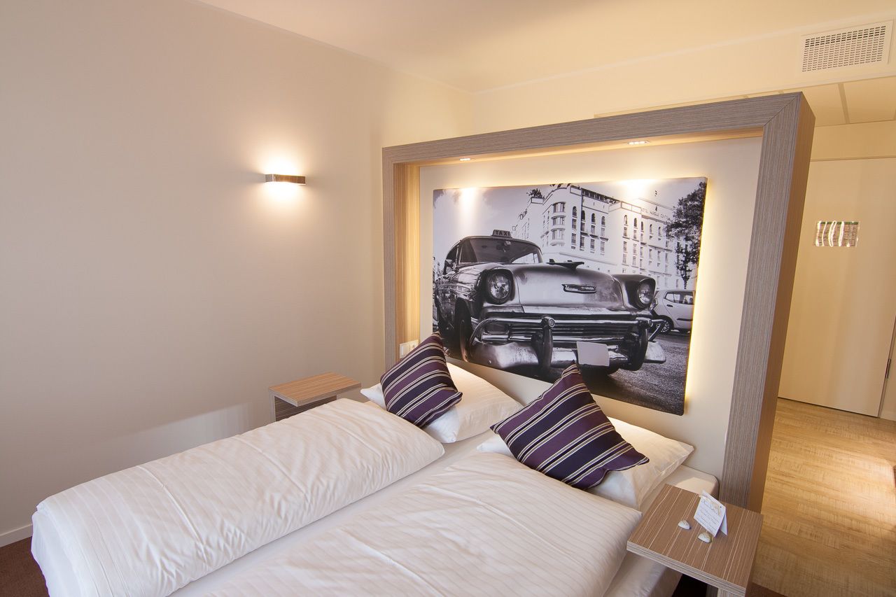 Een hotelkamer met een groot bed, twee kussens, een nachtkastje, een lijst met een afbeelding aan de muur, en een klein tafeltje met een bordje.