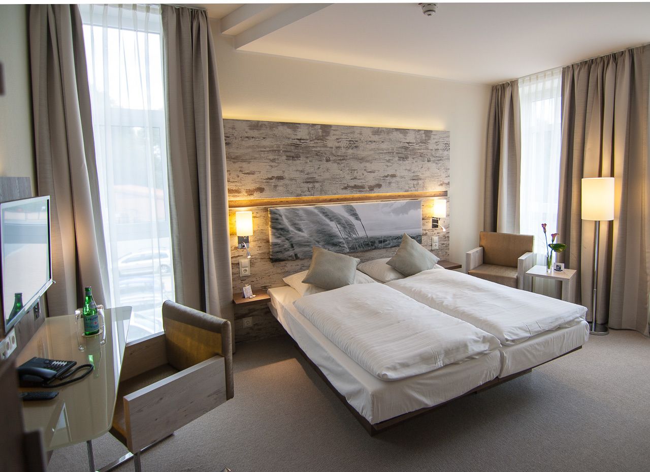 Una habitación de hotel con una cama grande, un escritorio con computadora, una silla y una mesita de noche con lámpara.