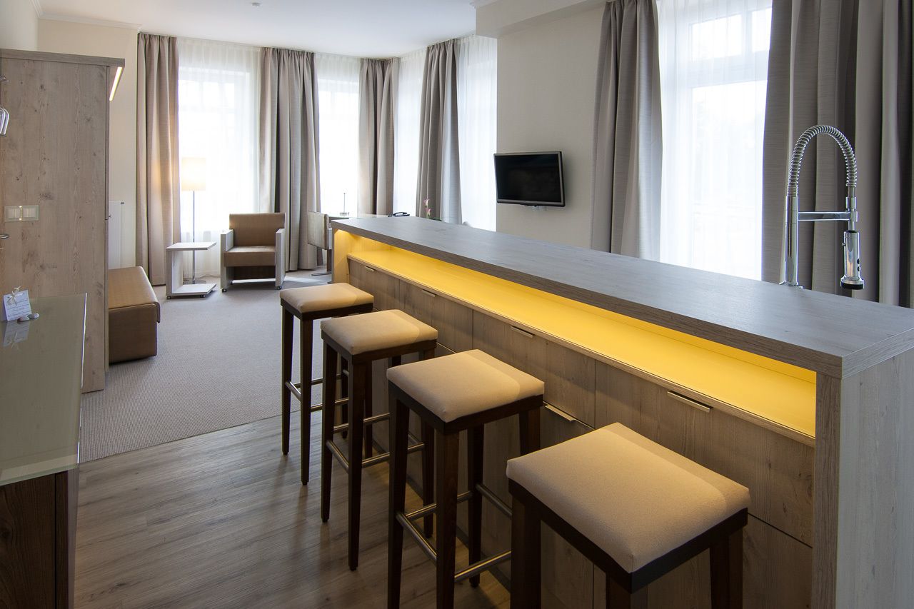 Une chambre d’hôtel moderne dotée d’un long bar, de quatre tabourets de bar, d’un téléviseur à écran plat et d’une grande fenêtre avec des rideaux.