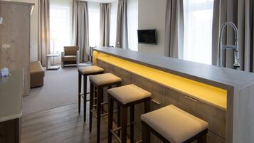 Une chambre d’hôtel moderne dotée d’un long bar, de quatre tabourets de bar, d’un téléviseur à écran plat et d’une grande fenêtre avec des rideaux.